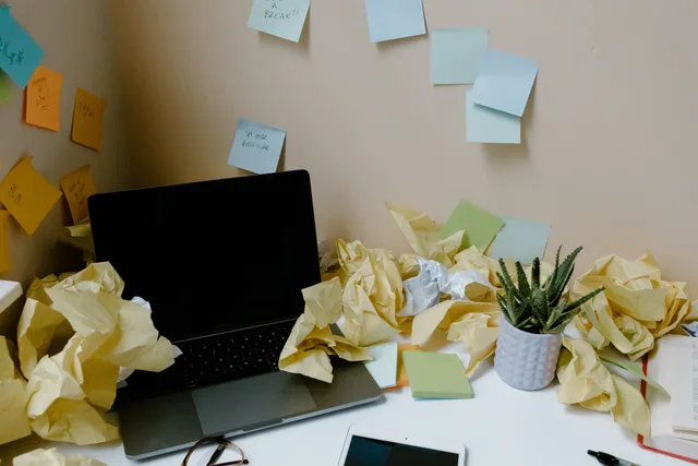 A chaotic office desk with a laptop, scattered papers, sticky notes, and a potted plant. Avtalshantering för småföretag: från offert till underskrift